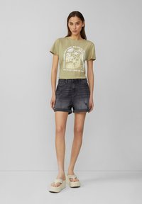 T-shirt court vert olive avec impression graphique blanche, associé à un short en jean noir taille haute et des sandales à plateforme claires.