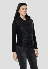 Črna jakna s kapuco, spredaj z rjušasto podlogo, z zadrgo in stranskimi žepi. Iz mehke tkanine z gladko teksturo.