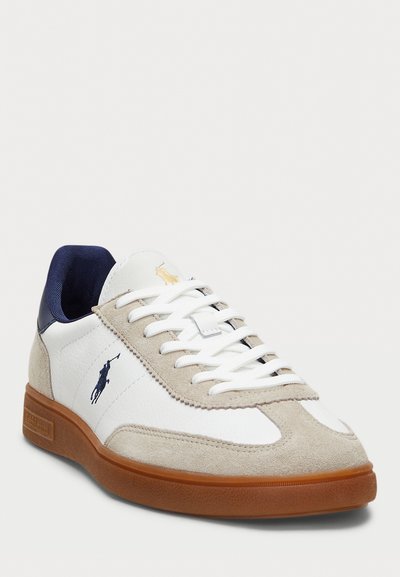 Zapatilla blanca con acentos de ante gris, talón azul marino y suela de goma. Presenta cordones y un logo dorado en el costado. Diseño atlético clásico.