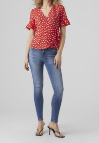 Blusa avvolgente rossa a fiori con maniche corte a volant, abbinata a jeans skinny blu e sandali neri con tacco. Texture del tessuto liscia.