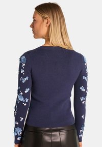 Marineblaue Strickjacke mit langen Ärmeln, die 3D-Blumenapplikationen in Hellblau und Weiß an den Ärmeln aufweist. Glatte Textur, Rundhalsausschnitt.