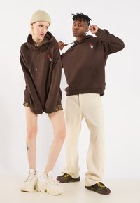 Bruine capuchontruien met een voorkraag en een klein hartontwerp. Model draagt een beige beanie, de ander heeft beige broeken en sneakers aan.