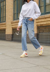 Gray cropped hoodie, light blue wide-leg jeans, and multicolored sneakers with pink and orange accents. Person holds pink sunglasses.