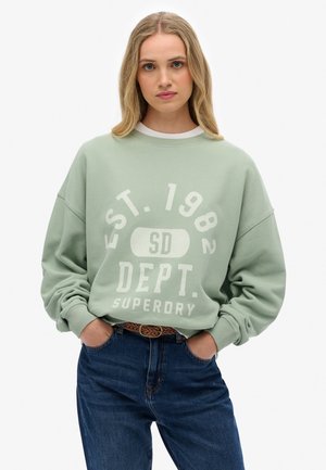 Muntgroene sweatshirt met witte graphic tekst, ronde halslijn en verlaagde schouders, gecombineerd met donkerblauwe jeans en een geweven riem.