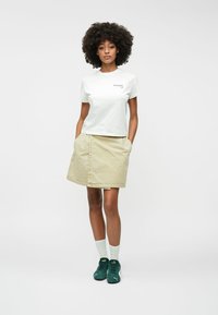 Camiseta corta blanca con logo, falda de mezclilla beige con bolsillos y zapatillas verdes con calcetines blancos. La modelo está de pie con las manos en los bolsillos.