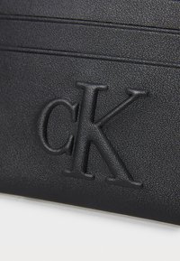Portafoglio in pelle nera testurizzata con logo "ck" in rilievo nell'angolo in basso e dettagli di cucitura orizzontale.