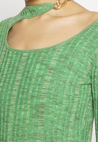 Maglione verde a costine con un collo a taglio unico, accentuato da un anello dorato e leggere variazioni di texture su tutto il tessuto.