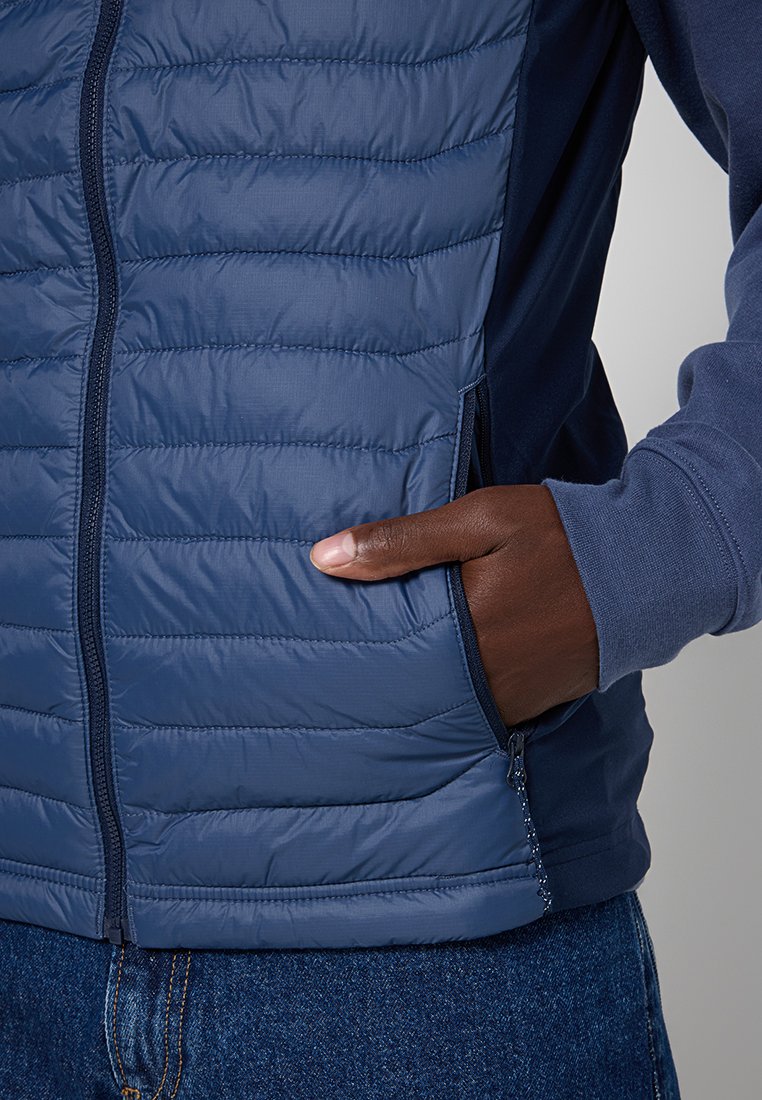 Mano nella tasca di un gilet blu trapuntato con zip indossato sopra un felpa blu coordinato e jeans blu.