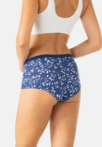 Femme portant des sous-vêtements imprimés floraux bleus et un crop top blanc, vue de dos sur fond blanc.