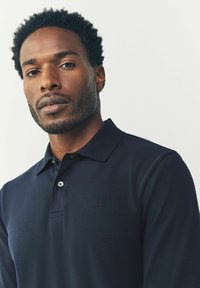 Camiseta polo en color navy que presenta un tejido texturizado, tapeta de botones y un pequeño logo en el pecho. Con mangas cortas y un diseño de cuello clásico.