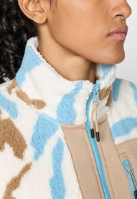 Primo piano di una persona che indossa una giacca in pile con un collo alto, caratterizzata da motivi astratti blu, bianchi e beige e una zip blu.