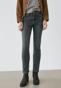 Slim-fit mörkgrå jeans kombinerade med en brun jacka, grå topp och svarta mockaankelboots mot en enfärgad bakgrund.