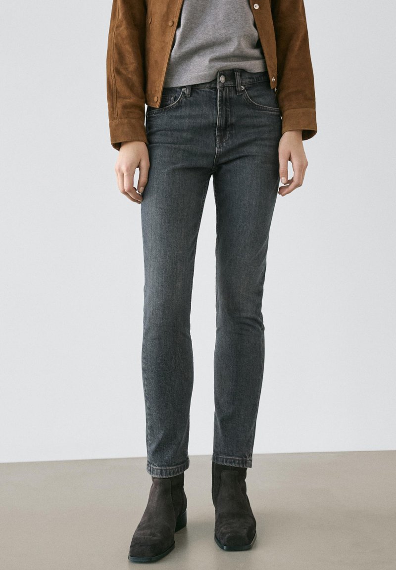 Slim-fit mörkgrå jeans kombinerade med en brun jacka, grå topp och svarta mockaankelboots mot en enfärgad bakgrund.