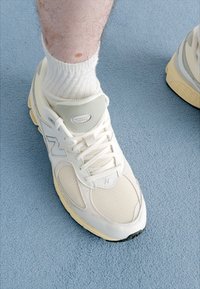 Weiße New Balance Sneaker mit Mesh- und Lederdetails, hellgrauen Akzenten, ausgestattet mit einer gepolsterten Sohle und geschichtetem Design, kombiniert mit einer weißen Socke.