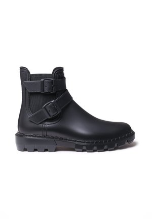 Zwarte enkelboot gemaakt van glad rubber met twee gespsluitingen, gestructureerde bovenkant, versterkte stiksels en een robuuste zool met grippatroon.