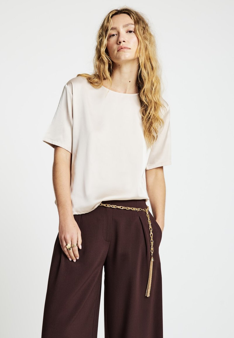 Top beige a maniche corte, tessuto liscio, vestibilità ampia. Abbinato a pantaloni a gamba larga color marrone scuro e cintura a catena dorata con dettaglio a nappina.