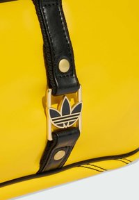 Žuta sintetička torba s crnim remenom i zlatnom kopčom. Istaknut Adidasov logo u crnoj boji, naglašavajući sportski, suvremen dizajn.
