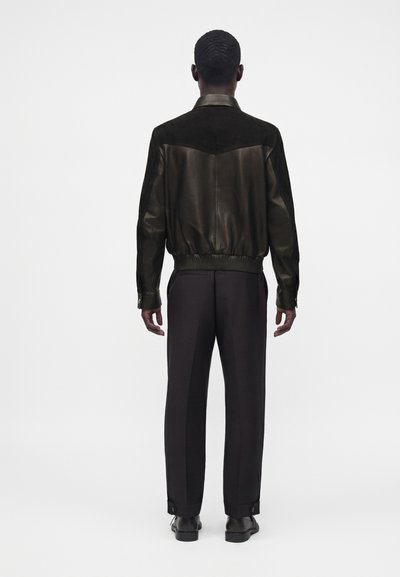 Homme portant une veste en cuir et suède noire, un pantalon noir et des chaussures noires, debout, de dos, sur un fond blanc uni.