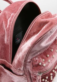 Rucksack aus pinkfarbenem Samt mit einem Reißverschluss-Öffnung, strukturierten Oberflächen, Perlenverzierungen und einem schwarzen Innenraum mit sichtbarem Markenetikett.