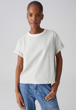 Femme portant un t-shirt à manches courtes rayé blanc et gris clair et un jean bleu taille haute avec des poches plaquées, debout devant un fond uni.