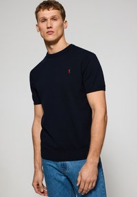 Mörkblå kortärmad tröja i stickad tyg, med rund halsringning och en liten röd logotyp på bröstet.