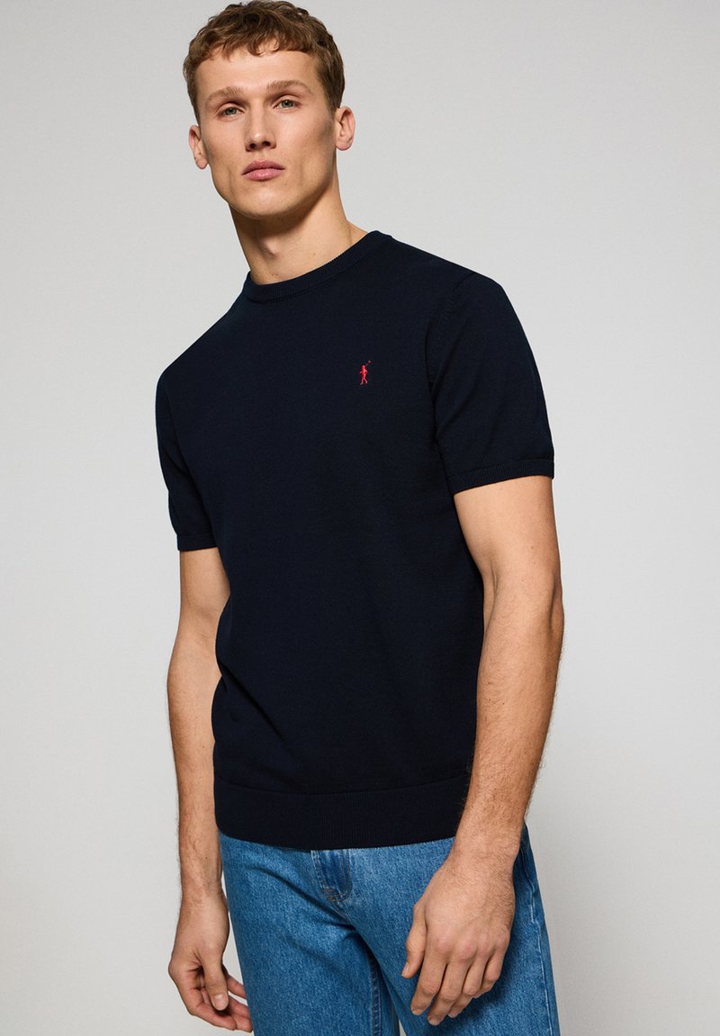 Mörkblå kortärmad tröja i stickad tyg, med rund halsringning och en liten röd logotyp på bröstet.