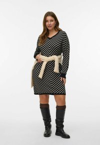 Robe rayée noire et blanche avec un décolleté en V, des manches longues et une taille cintrée à l'aide d'une ceinture en tricot couleur crème. Le modèle porte des bottes noires jusqu'aux genoux.