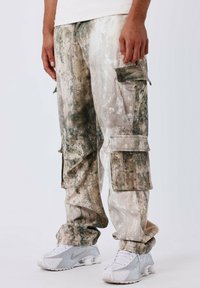 Cargo pants in een vervaagd beige en groen patroon, met meerdere zakken en een losse pasvorm. Gecombineerd met lichtgrijze sportschoenen.