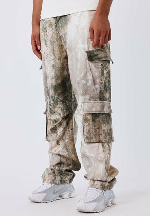 Cargo pants in een vervaagd beige en groen patroon, met meerdere zakken en een losse pasvorm. Gecombineerd met lichtgrijze sportschoenen.
