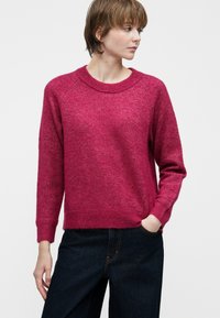 Młoda osoba z krótkimi brązowymi włosami, ubrana w różową bluzę z długim rękawem i ciemnoniebieskie dżinsy, patrzy w bok z jedną ręką w kieszeni.