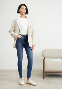 Blazer de lino beige claro sobre una blusa blanca, combinado con jeans de mezclilla azul ajustados y zapatillas blancas con suelas beige. Diseño sencillo y moderno.