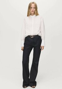 Witte lange mouwen blouse met plooien aan de voorkant, gecombineerd met donkere flared jeans en een bruine riem met een ronde metalen gesp.