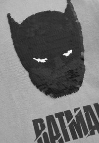 Graues T-Shirt mit einem Design eines schwarzen Pailletten-Batman-Masken. Die Pailletten erzeugen eine strukturierte Oberfläche, mit weißen Augen und einem auffälligen Text darunter.