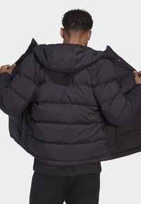 Schwarze Daunenjacke mit Kapuze, ausgestattet mit einem gesteppten Design, Reißverschluss und zwei seitlichen Taschen, wird von hinten offen getragen.