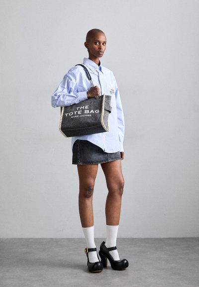 Sac fourre-tout gris avec des accents crème et un grand texte blanc, porté sur une chemise bleue et une jupe noire, associé à des chaussettes blanches et des talons noirs.
