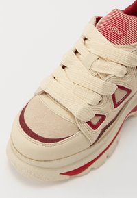 Zapatilla beige con parte superior texturizada, acentos rojos y cordones color crema. Presenta una suela gruesa y cortes triangulares en los laterales.
