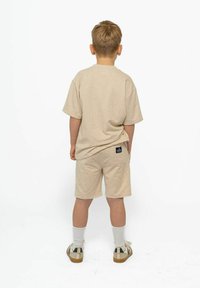 Beige kokvilnas T-krekls un šortu komplekts, brīvas formas, īsas piedurknes, aizmugurējā kabata šortos, kopā ar baltām zeķēm un sporta apaviem.