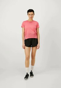 Kvinde står iført en lyserød kortærmet skjorte, sorte shorts, hvide sokker og sorte sneakers foran en ensfarvet lys baggrund.