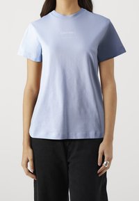 Světle modré bavlněné tričko s krátkými rukávy, kulatým výstřihem a bílým logem "Calvin Klein" na přední straně. Hladká textura, uvolněný střih.