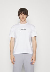 Witte katoenen t-shirt met korte mouwen, voorzien van een zwart "Calvin Klein" logo op de borst. Gepaarde met grijze sportbroeken.