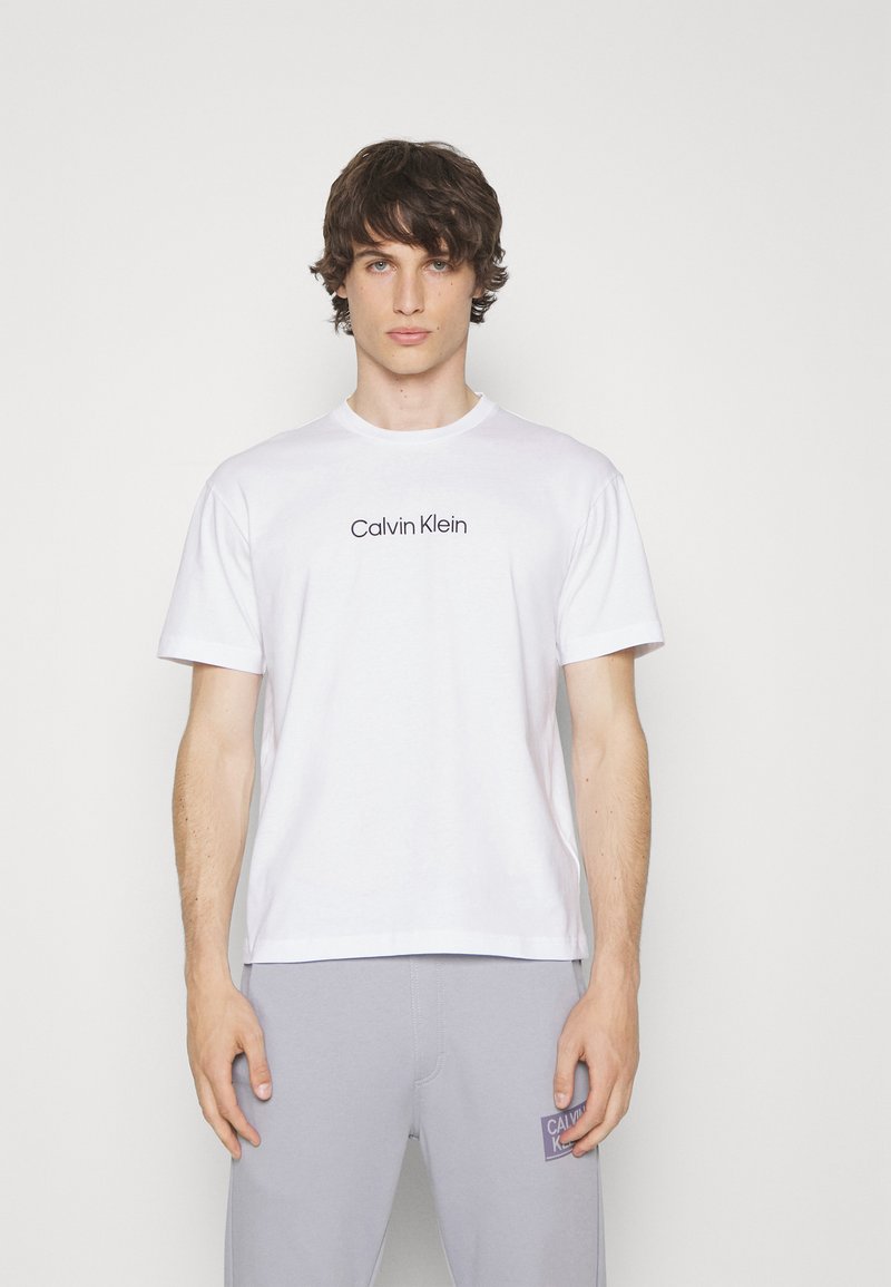 Witte katoenen t-shirt met korte mouwen, voorzien van een zwart "Calvin Klein" logo op de borst. Gepaarde met grijze sportbroeken.