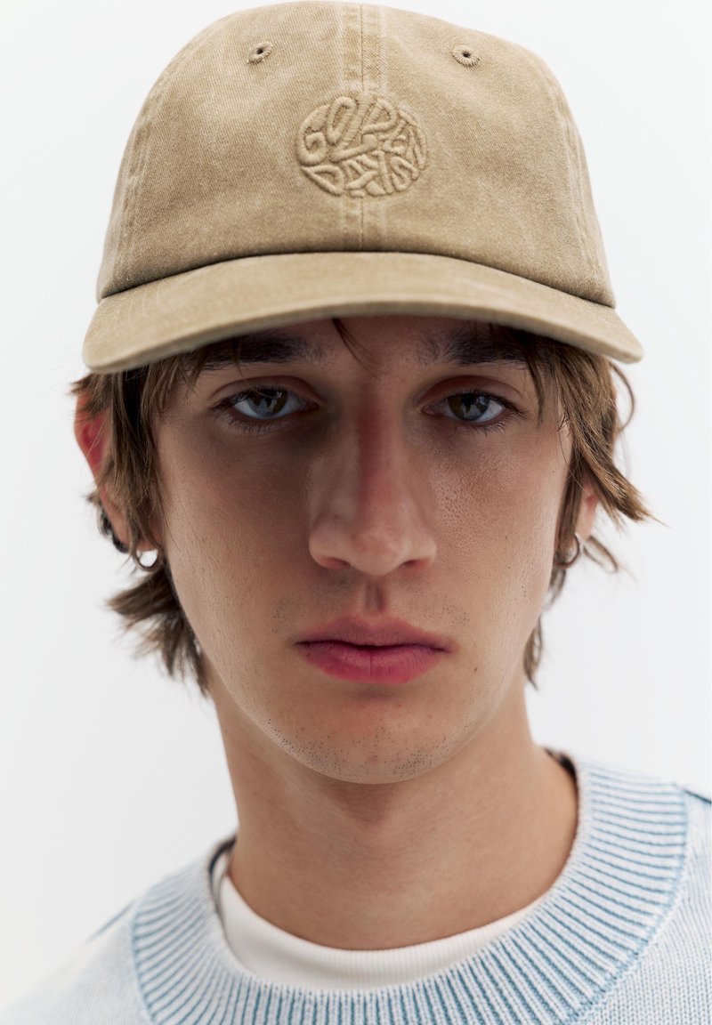 Jeune homme aux cheveux bruns porte une casquette beige avec un texte brodé et un pull bleu clair, regardant droit devant sur fond blanc.