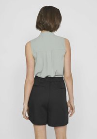 Hellgrüne ärmellose Bluse mit Faltendetail, kombiniert mit schwarzen Tailored-Shorts mit hinteren Taschen und einer glatten Stoffstruktur.
