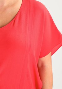 Blusa corallo brillante con scollo ampio e maniche corte, realizzata in un tessuto leggero e morbido con una sottile texture.