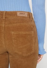 Hnědé sametové kalhoty s kapsou na zadní straně a koženou značkovou nálepkou, nošené s světle modrým žebrovaným dlouhým rukávem, ruka částečně viditelná.