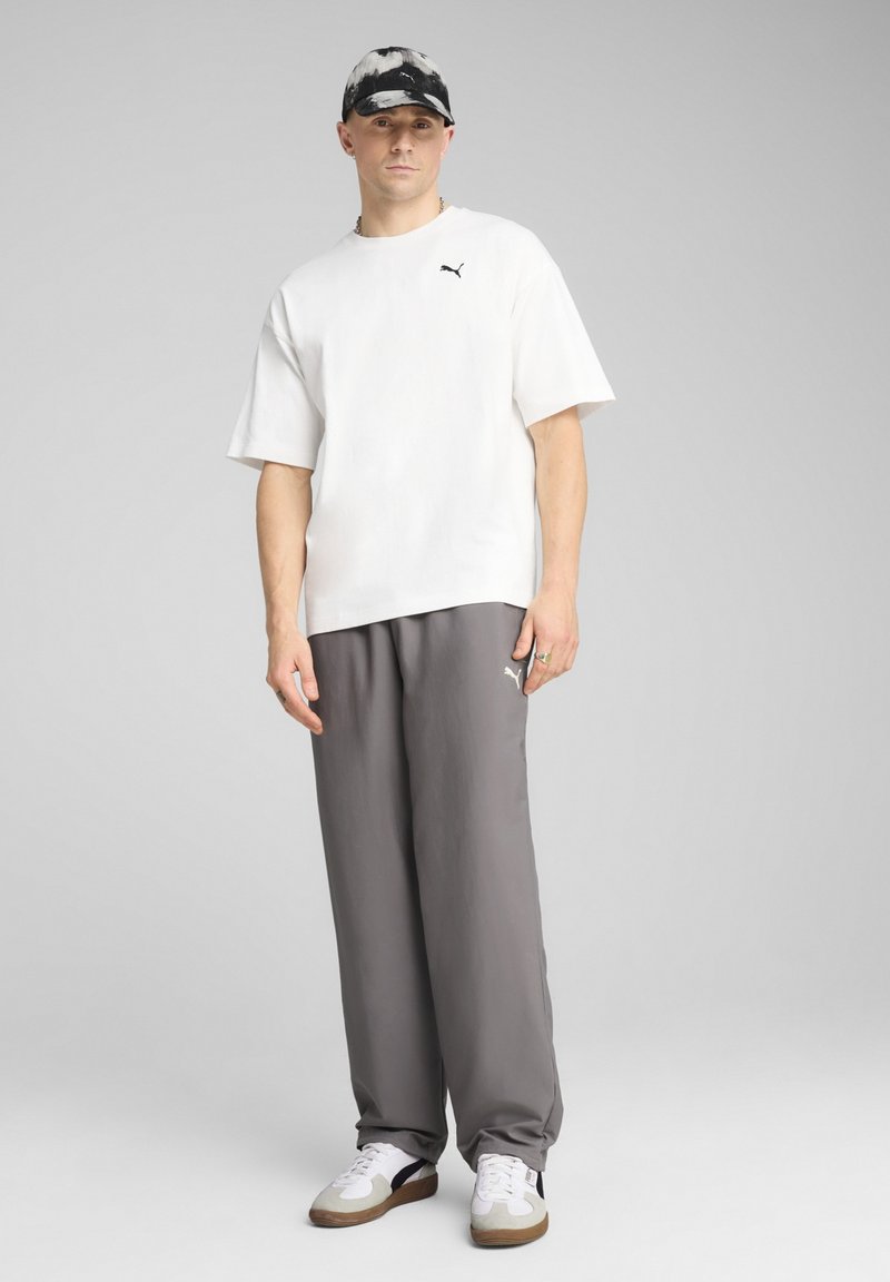 Camisa blanca de manga corta con logo negro, pantalones grises de pierna ancha y zapatillas blancas. El accesorio para la cabeza tiene un patrón de tinte anudado en negro y gris.