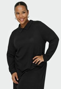 Černé dlouhé šaty s límečkem, hladkou texturou a knoflíkovými manžetami. Modelka pózuje s rukou na boku a ukazuje střih oděvu.