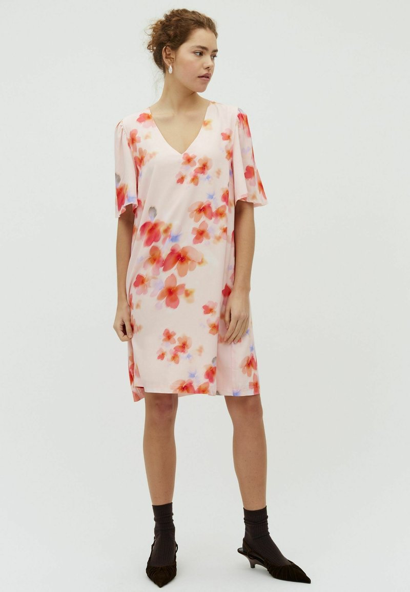 Femme portant une robe fleurie rose pâle jusqu'aux genoux avec des manches courtes, associée à des chaussettes noires et des chaussures pointues noires.