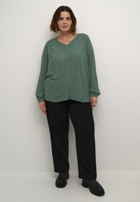 Haut vert à manches longues avec un col en V et un tissu texturé, associé à un pantalon noir à jambes larges et des bottes noires chunky.