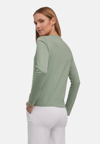 Langarmshirt mit Rundhalsausschnitt in Hellgrün, aus weichem Stoff gefertigt. Die Rückansicht zeigt einen taillierten Schnitt und eine glatte Textur, kombiniert mit weißen Hosen.